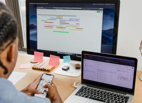 Businessman multitasks with smartphone, laptop, and monitor in office setting.